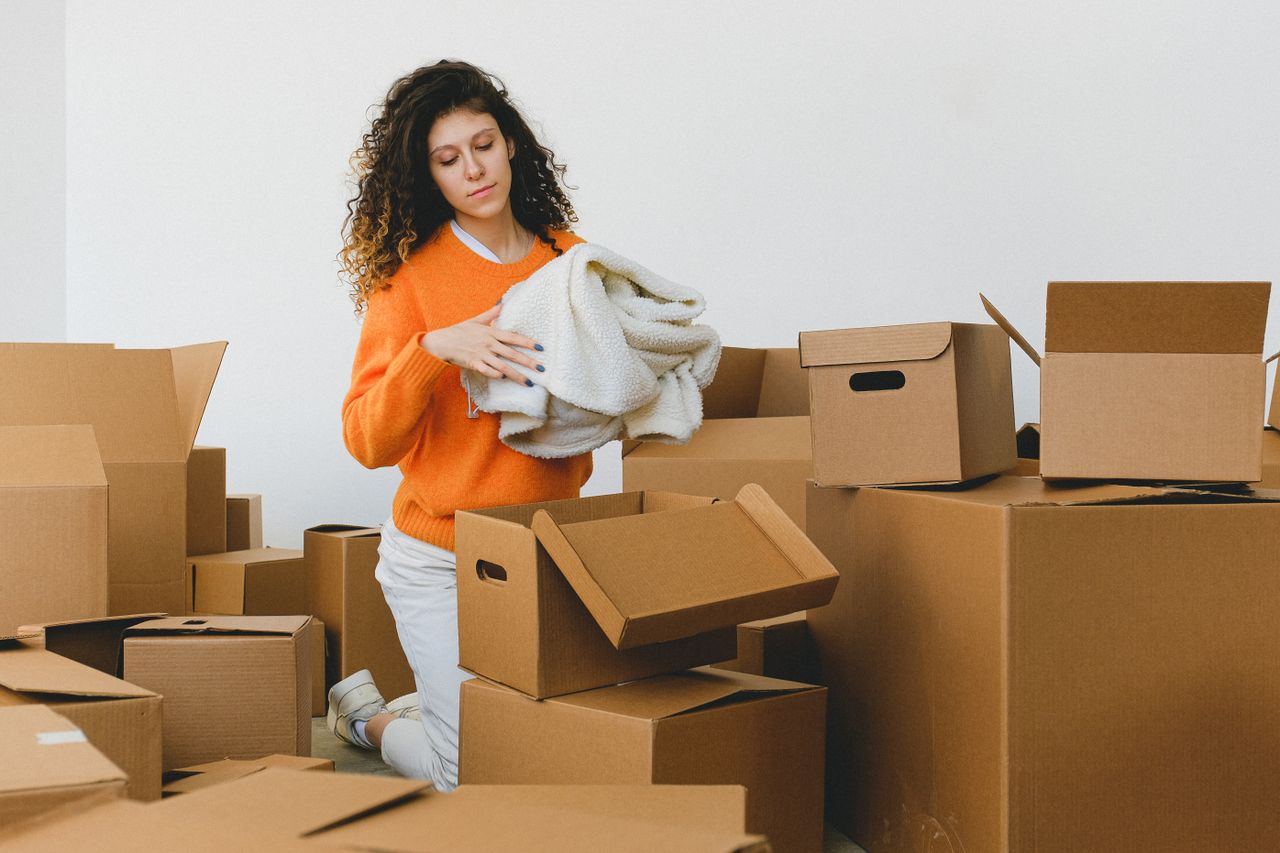 our-story Concentrated young lady with curly hair standing on knees on floor and packing clothes into carton boxes before moving into new flat