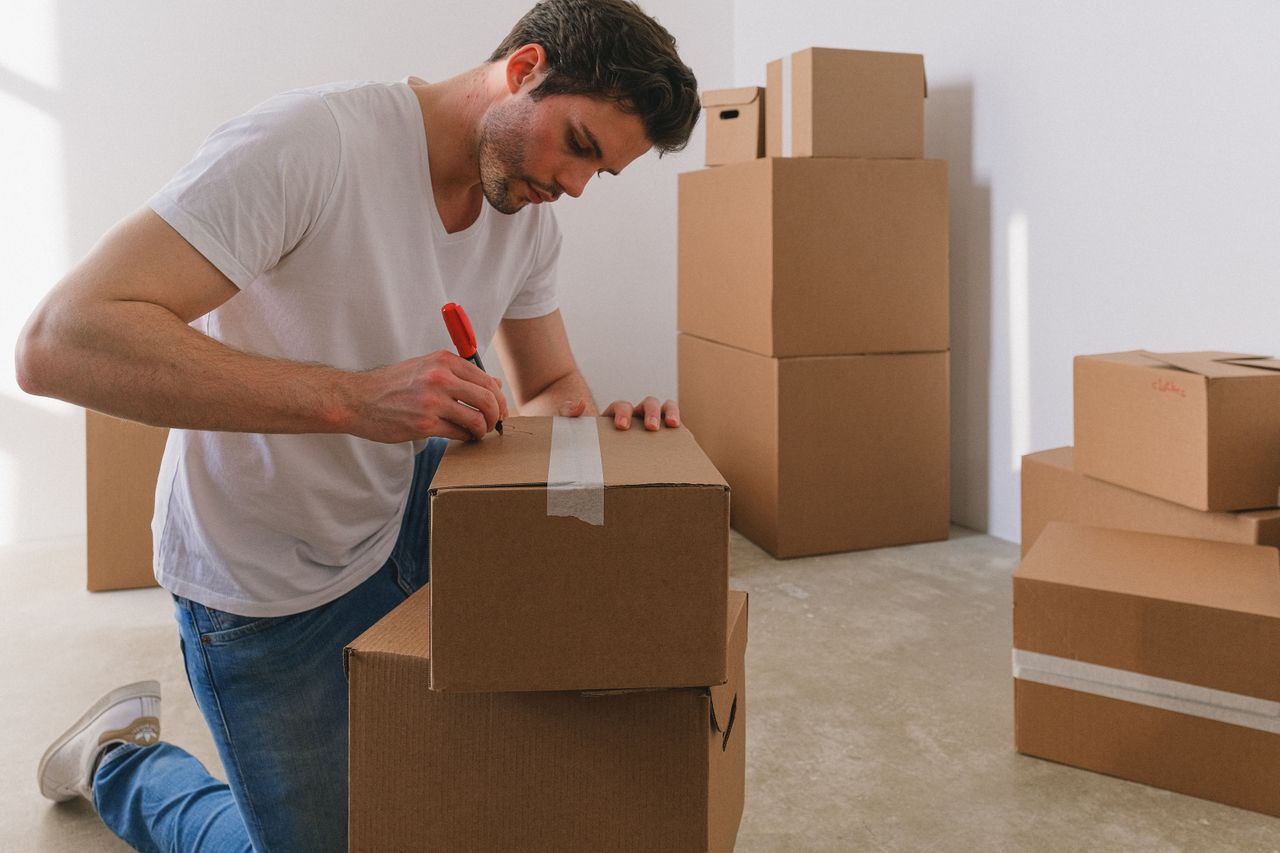 A man labels cardboard boxes for moving, kneeling indoors with natural light.